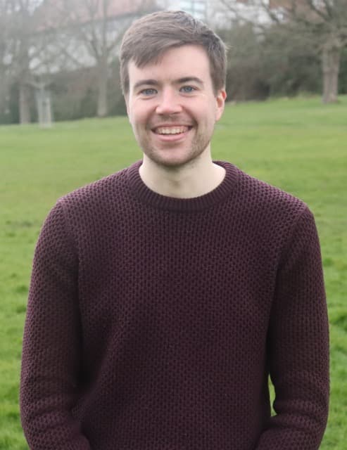 Nick Humberstone in a burgundy knitwear jumper, standing in a park with green grass and a treeline in the background.